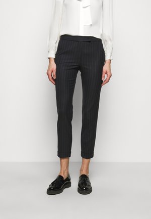 Woman wearing black pinstripe cropped trousers, white long-sleeve blouse, and black shiny tassel loafers, standing against a plain background.