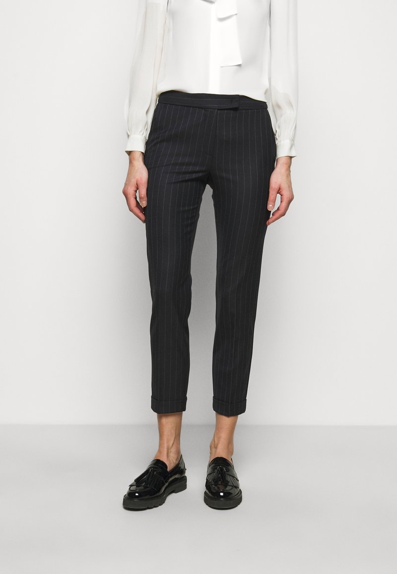 Mujer con pantalones cortos negros a rayas, blusa blanca de manga larga y mocasines negros brillantes con borlas, posando frente a un fondo liso.