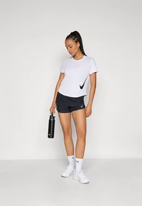 Hvit Nike T-skjorte med svart swoosh, kombinert med svarte shorts. Modellen holder en svart vannflaske, iført hvite sokker og joggesko.