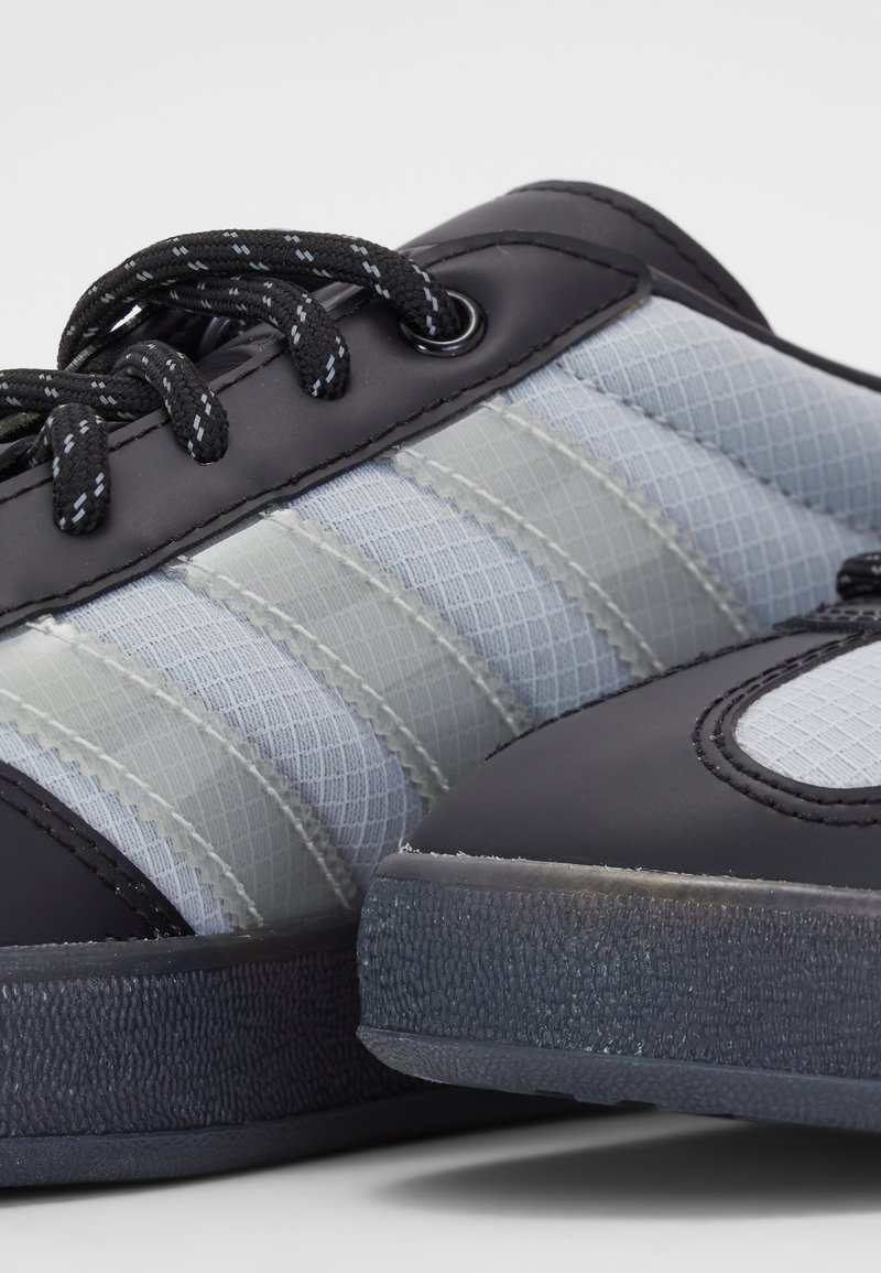 Schwarze Sneakers mit einem synthetischen Obermaterial, das graue Textileinsätze und eine strukturierte schwarze Gummisohle aufweist. Schwarze Schnürsenkel und Ösen vervollständigen das Design.