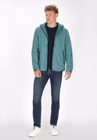 Jachetă ușoară cu glugă în nuanța teal, cu un model geometric, asortată cu blugi închiși și adidași albi. Jacheta are buzunare cu fermoar.