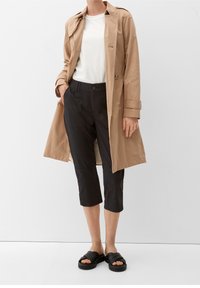 Donna che indossa un trench beige sopra una camicia bianca, pantaloni neri cropped e sandali neri imbottiti, in piedi contro un muro bianco.