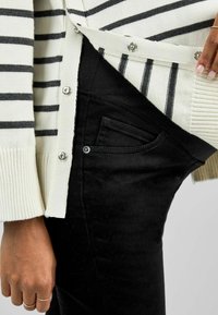 Gestreifter creme- und schwarzer Cardigan mit Druckknöpfen, über schwarzen Skinny Jeans aus strukturiertem Stoff mit dezenten Taschendetails getragen.