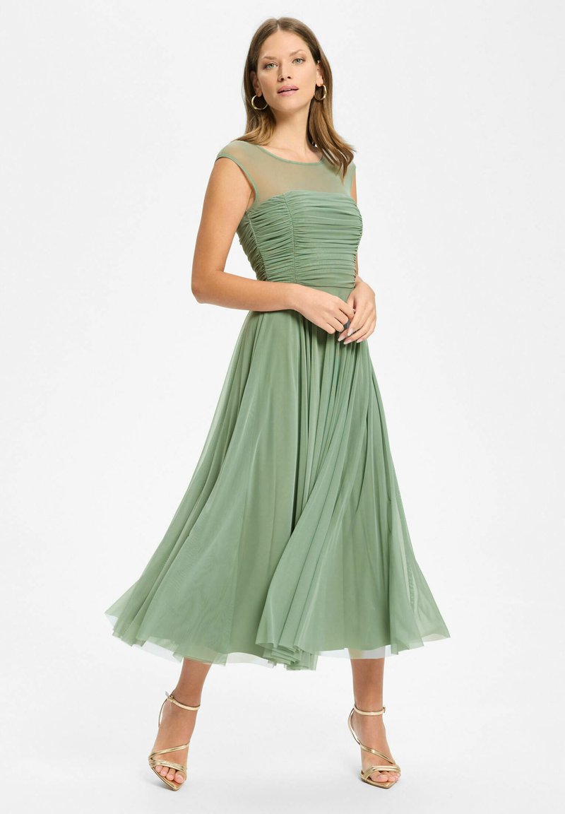 Femme portant une robe midi vert sauge sans manches avec un col transparent, associée à des talons dorés à brides, posant sur fond blanc.