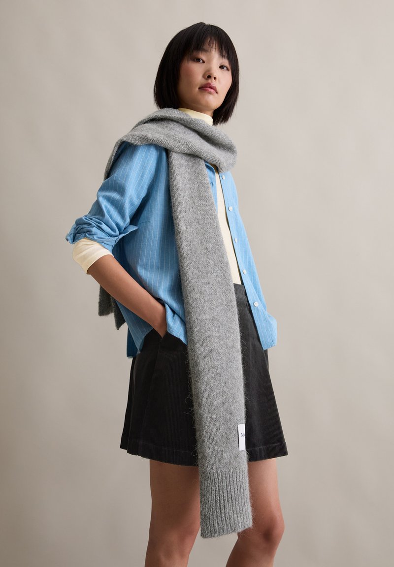 Grijze wollen sjaal gedrapeerd over een lichtblauw gestreept overhemd met knopen, gedragen met een zwarte corduroy rok; zachte textuur en ontspannen pasvorm.