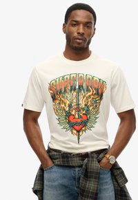 Camiseta crema con un gráfico colorido que presenta un corazón, una espada, llamas y alas. Tejido de algodón, de manga corta, corte relajado, combinada con jeans.