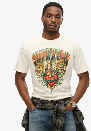 T-shirt crème avec un graphisme coloré représentant un cœur, une épée, des flammes et des ailes. Tissu en coton, manches courtes, coupe décontractée, porté avec un jean.