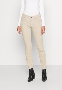 Jeans skinny beige con una leggera lucentezza, dotati di tasche frontali e orli arrotolati, abbinati a stivali neri alla caviglia. Texture liscia.