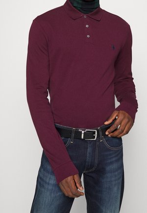 Homme portant un polo bordeaux à manches longues par-dessus un col roulé à carreaux foncé, un jean foncé et une ceinture noire avec une boucle argentée.