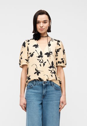 Femme portant un chemisier beige à imprimé floral noir avec des manches bouffantes, associé à un jean taille haute bleu et un collier ras-de-cou noir à motif fleur.