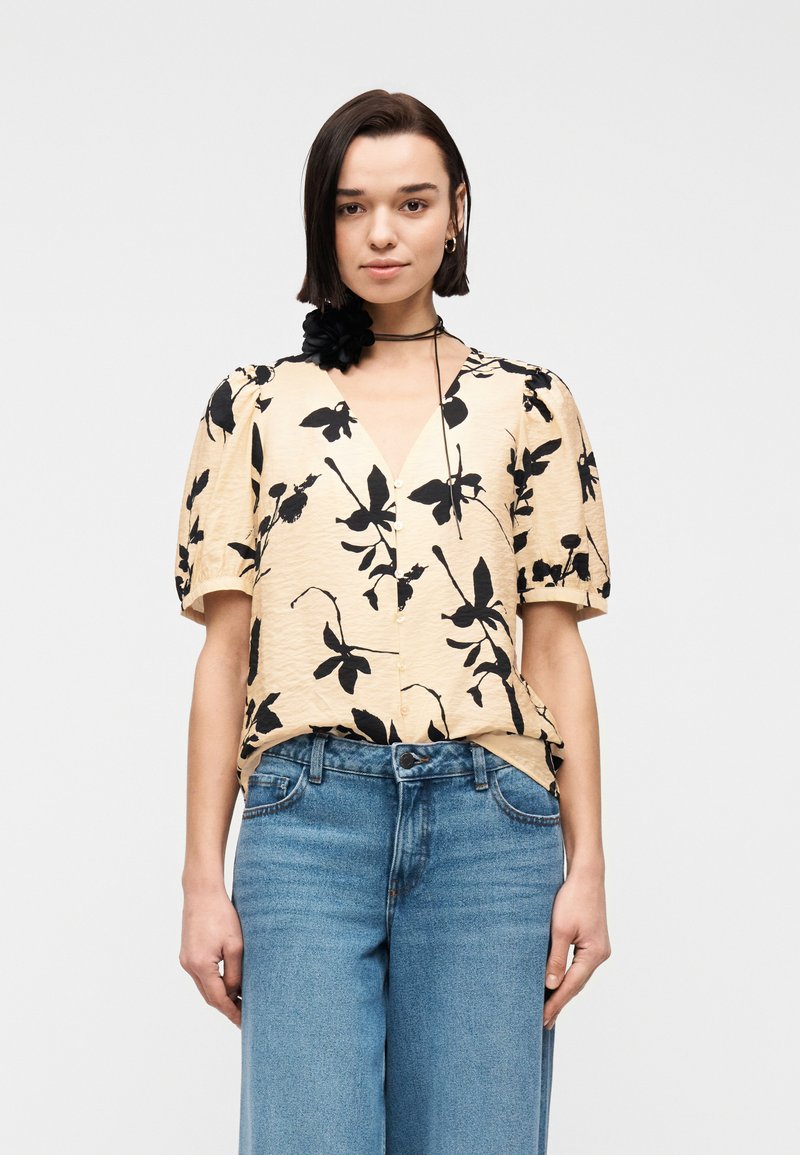 Vrouw draagt een beige blouse met zwart bloemenprint en pofmouwen, gecombineerd met een high-waisted blauwe jeans en een zwarte bloem choker.