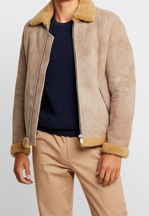 Hombre con chaqueta beige de borrego con cuello y puños esponjosos, suéter azul marino y pantalones beige, de pie frente a un fondo neutro.