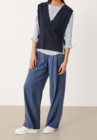 Top in maglia senza maniche di colore blu navy sopra una maglietta a maniche lunghe a righe, abbinato a pantaloni larghi blu chiaro e sneaker bianche. Design semplice.