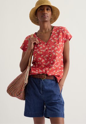 Vrouw met een strohoed, een rood bloemenblouse met korte mouwen, spijkershort, een bruine riem en een meerkleurige gewebde boodschappentas op haar schouder.
