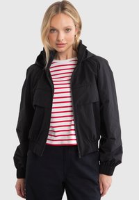 Black cropped jacket with hood, elastic cuffs, and two front pockets. Worn over a red and white striped shirt. Smooth, lightweight fabric.