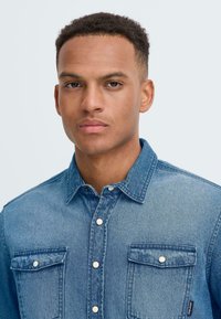 Denim shirt in light blue with a button-down collar, two chest pockets, and silver snap buttons. Texture appears smooth and slightly faded.