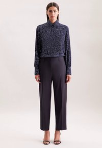 Blusa blu navy con pois bianchi, con collo e maniche lunghe, abbinata a pantaloni scuri su misura e tacchi neri aperti sulle dita.