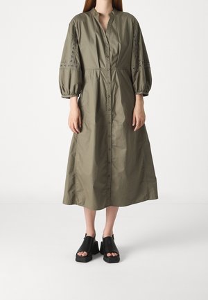 Olivgrünes, knopfgeschlossenes Kleid mit hohem Kragen, Ballonärmeln und bestickten Details. Kombiniert mit schwarzen Plateau-Sandalen.