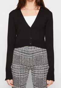 Czarny skrócony kardigan z dekoltem w serek, z przodu zapinany na guziki i z długimi rękawami. W połączeniu z spodniami w szaro-czarny wzór w jodły.