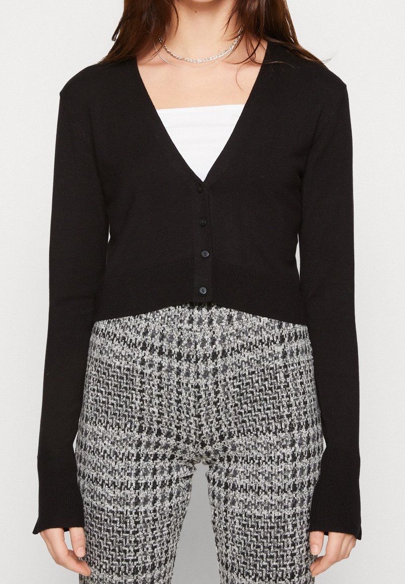 Czarny skrócony kardigan z dekoltem w serek, z przodu zapinany na guziki i z długimi rękawami. W połączeniu z spodniami w szaro-czarny wzór w jodły.
