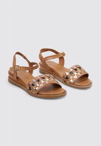 Sandalias marrones con punta abierta, que presentan un diseño de tira trenzada en negro y acentos metálicos. Tira delgada en el tobillo con hebilla y suela plana.
