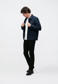 Fiatal férfi, aki egy sötétkék zakót igazgat fehér ing fölött, fekete nadrágot és fekete fűzős csizmát visel, egyszínű fehér háttér előtt áll.