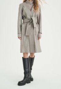 Jasnoszary skórzany płaszcz o obcisłym kroju, z paskiem w talii, dwiema przednimi kieszeniami i długimi rękawami, zestawiony z dopasowanymi czarnymi kozakami do kolan.