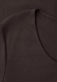 Nærbillede af mørkebrunt stof med synlige syninger og en buet søm, sandsynligvis en del af et tøjs halsudskæring og skulderområde.