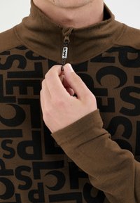 Die Hand einer Person zieht den Reißverschluss eines braun-schwarz gemusterten Langarmoberteils mit hochgeschlossenem Kragen und einem "SLOPE"-Tag am Reißverschluss.