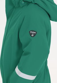 Lys grøn vandtæt jakke med hætte, der har et sort "ZIGZAG" logo-patch på armen og en hvid reflekterende stripe på ærmet.