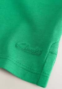Tissu en coton vert avec une texture fine, arborant un logo "Clarks" brodé. Le vêtement présente un détail de bordure cousue soigné.