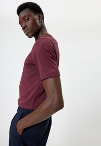 T-shirt a maniche corte bordeaux realizzato in tessuto morbido, con scollo rotondo e cuciture pulite, abbinato a pantaloni navy, posato di lato.