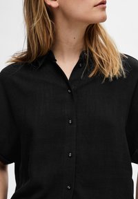 Chemise noire à boutons fabriquée en tissu texturé, dotée d'un col et de manches courtes, avec de subtils boutons sur le devant.