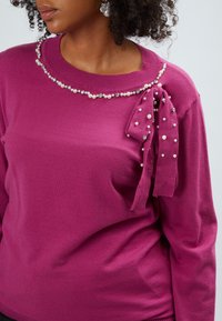 Maglione fucsia con scollatura rotonda, impreziosito da accenti di perle e un dettaglio di fiocco arricciato sulla spalla sinistra. Texture morbida e lavorata a maglia.