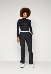 Černý sportovní top s dlouhým rukávem a vysokým límcem, doplněný černými rozšířenými kalhotami a bílým opaskem. Modelka má na nohou bílé tenisky.