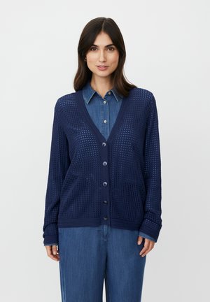 Cárdigan de punto azul marino con un diseño de malla, que presenta un escote en V, botones frontales y mangas largas. Usado sobre una camisa de mezclilla.