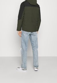 Grön och svart isolerad jacka med huva, försedda med en logotyp på ärmen, i kombination med ljusblå slitna jeans och grå sneakers.