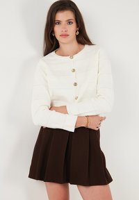 Weißer strukturierter Cardigan mit langen Ärmeln, fünf goldenen Knöpfen, kombiniert mit einem dunkelbraunen Faltenrock.