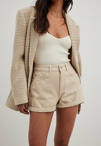 Khaki denim shorts med oprullede kanter, parret med en cremefarvet ribbet tank top og en struktureret beige blazer. Materialerne er bløde og robuste.