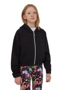 Cappotto corto nero con zip frontale e due tasche, abbinato a leggings con motivo splatter colorato in rosa, verde e blu.