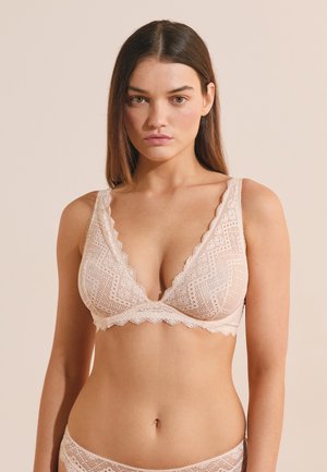 Kobieta o prostych brązowych włosach, ubrana w jasno różowy koronkowy bralette i pasujące koronkowe majtki na beżowym tle.