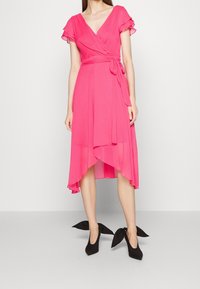 Robe portefeuille rose avec manches volantes et ourlet asymétrique. En tissu léger, nouée à la taille, assortie à des chaussures à talons noires.