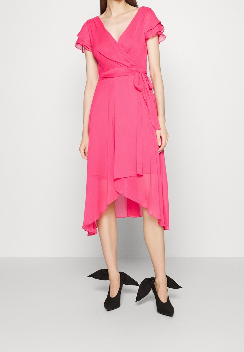 Robe portefeuille rose avec manches volantes et ourlet asymétrique. En tissu léger, nouée à la taille, assortie à des chaussures à talons noires.