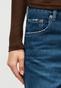 Χέρι δίπλα σε μπλε τζιν τζιν με μικρό φθαρμένο τσεπάκι για κέρματα, ορατό φθαρμένο καφέ ριγωτό μακρυμάνικο πάνω μέρος στο χέρι.