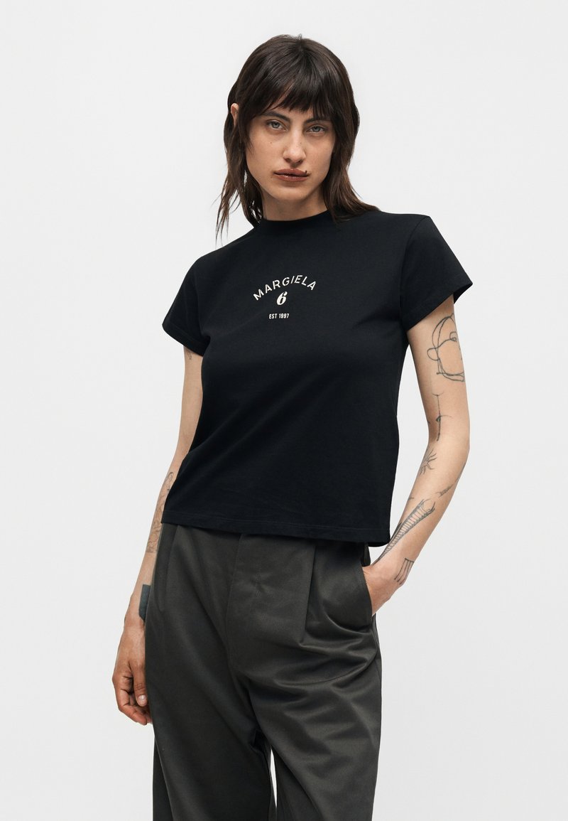 Woman with dark hair wearing a black Margiela t-shirt and dark pants, standing with one hand in pocket against a plain background.