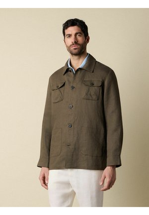 Uomo che indossa una giacca marrone con bottoni e tasche con patta sopra una camicia azzurro chiaro e pantaloni bianchi, in piedi di fronte a uno sfondo beige.