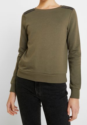 Nainen, joka pukeutunut oliivinvihreään pitkähihainen toppiin, jossa mustat helmikoristeiset olkapäät, ja mustiin farkkuihin, seisomassa yksinkertaista taustaa vasten.