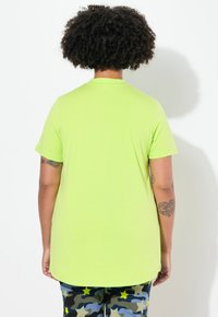Grøn T-shirt med korte ærmer og rund halsudskæring, glat tekstur og en længere bagkant, parret med bukser i camouflagemønster med gule stjerner.