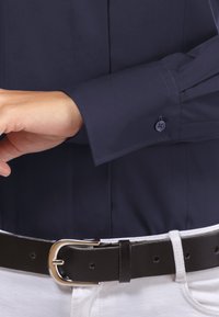 Dunkelblaues Hemd mit Knöpfen, figurbetontem Design, langen Manschetten und Nahtdetails, getragen mit einem schwarzen Ledergürtel und weißen Hosen.