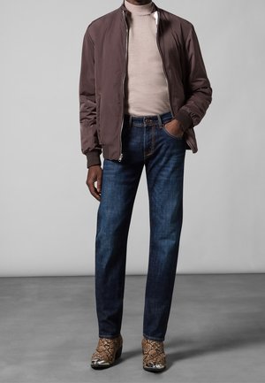 Chaqueta bomber marrón sobre un cuello de tortuga beige, jeans azules y botines marrones con estampado y acentos metálicos. Textura suave en todo.